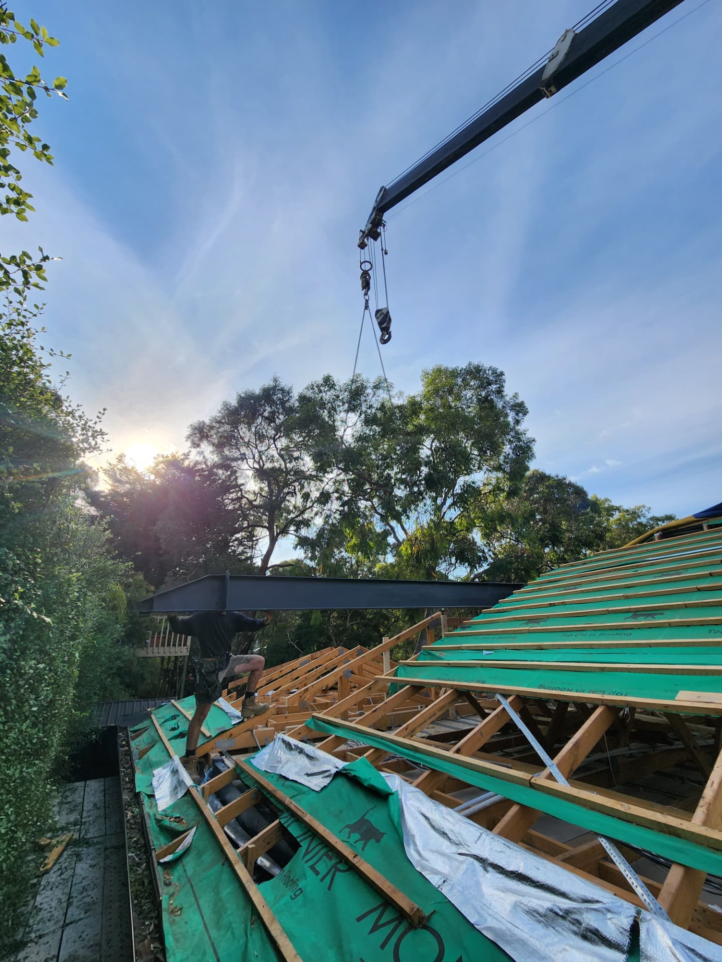 Crane lowering a steel beam onto a new timber roof frame at sunrise
